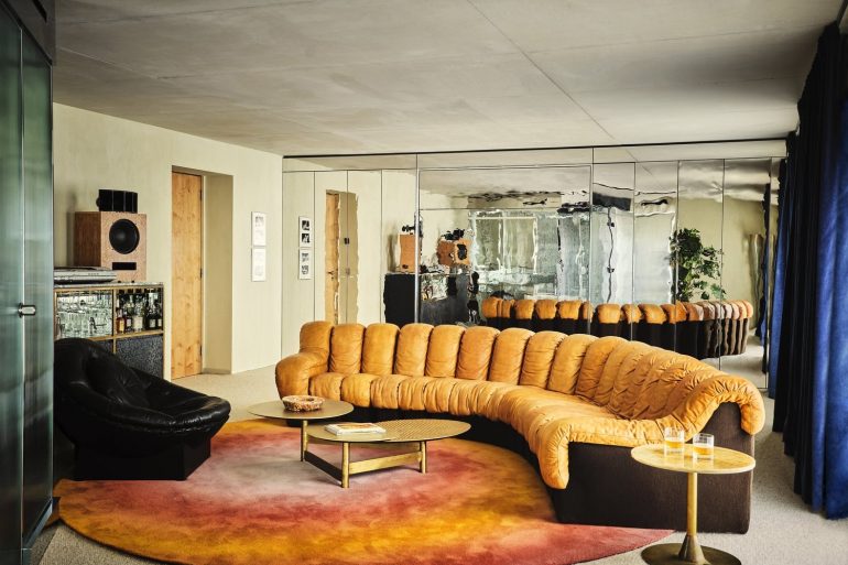 Mid‑century living room with a curved orange velvet sofa, black chair, and round coffee tables on a warm gradient rug by mirrored walls.