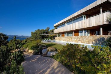Modern wooden-and-stone house with large glass windows, poolside terrace, and landscaped garden under a blue sky.