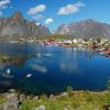 Colorful houses along a calm fjord with mountains in the background and their reflection in the water under a blue sky.