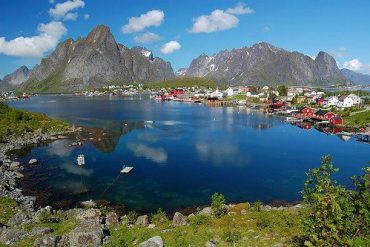 Colorful houses along a calm fjord with mountains in the background and their reflection in the water under a blue sky.
