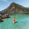 Woman in a black bikini stands waist-deep in crystal-clear turquoise water with a rugged green-covered rocky island in the background.