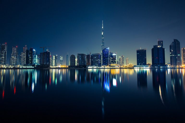 Night city skyline with illuminated skyscrapers reflecting on calm water.