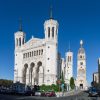 White ornate church facade with two tall spired towers and intricate carvings against a clear blue sky, street with parked cars in front.] ,