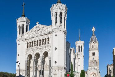 White ornate church facade with two tall spired towers and intricate carvings against a clear blue sky, street with parked cars in front.] ,