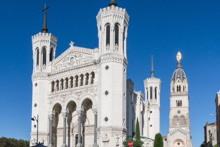 White ornate church facade with two tall spired towers and intricate carvings against a clear blue sky, street with parked cars in front.] ,