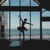 Silhouette of a ballet dancer in a tutu performing on a stage with a sea view through large windows behind her.