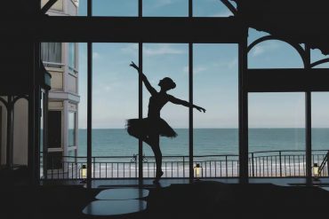 Silhouette of a ballet dancer in a tutu performing on a stage with a sea view through large windows behind her.
