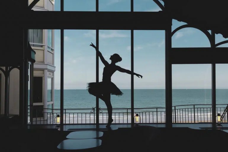 Silhouette of a ballet dancer in a tutu performing on a stage with a sea view through large windows behind her.
