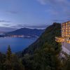Hotel perched on a cliff overlooking a lake and mountains at dusk, with warm-lit windows.