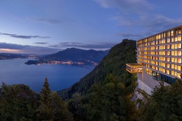 Hotel perched on a cliff overlooking a lake and mountains at dusk, with warm-lit windows.