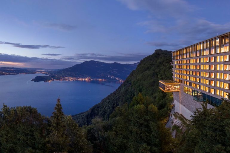 Hotel perched on a cliff overlooking a lake and mountains at dusk, with warm-lit windows.