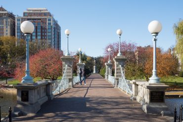 Pedestrian bridge with white globe street lamps, brick buildings, and autumn trees in the background.