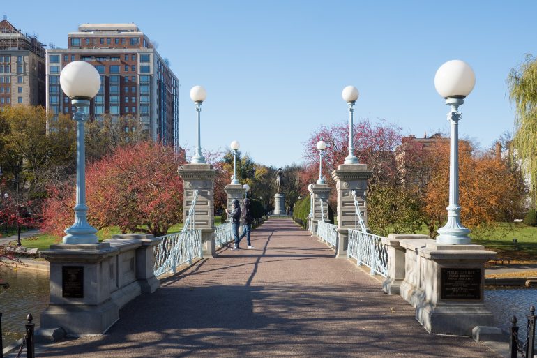 Pedestrian bridge with white globe street lamps, brick buildings, and autumn trees in the background.