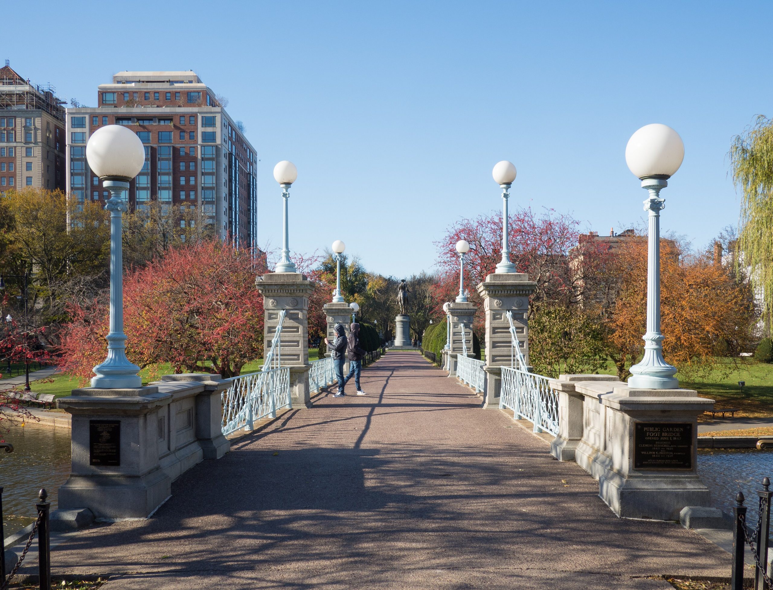 Pedestrian bridge with white globe street lamps, brick buildings, and autumn trees in the background.