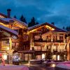 Snowy alpine lodge complex at dusk with warmly lit windows and a pink-lit decorative sculpture in front; tall Fir tree on the right lit with red lights.