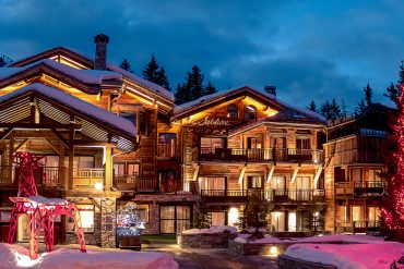 Snowy alpine lodge complex at dusk with warmly lit windows and a pink-lit decorative sculpture in front; tall Fir tree on the right lit with red lights.