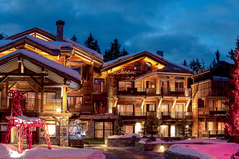 Snowy alpine lodge complex at dusk with warmly lit windows and a pink-lit decorative sculpture in front; tall Fir tree on the right lit with red lights.