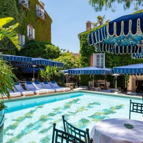 Poolside scene with turquoise water, white loungers under blue canopies and ivy-covered walls in a sunny resort setting.