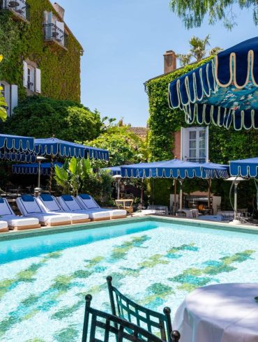 Poolside scene with turquoise water, white loungers under blue canopies and ivy-covered walls in a sunny resort setting.