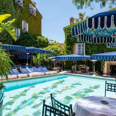 Poolside scene with turquoise water, white loungers under blue canopies and ivy-covered walls in a sunny resort setting.