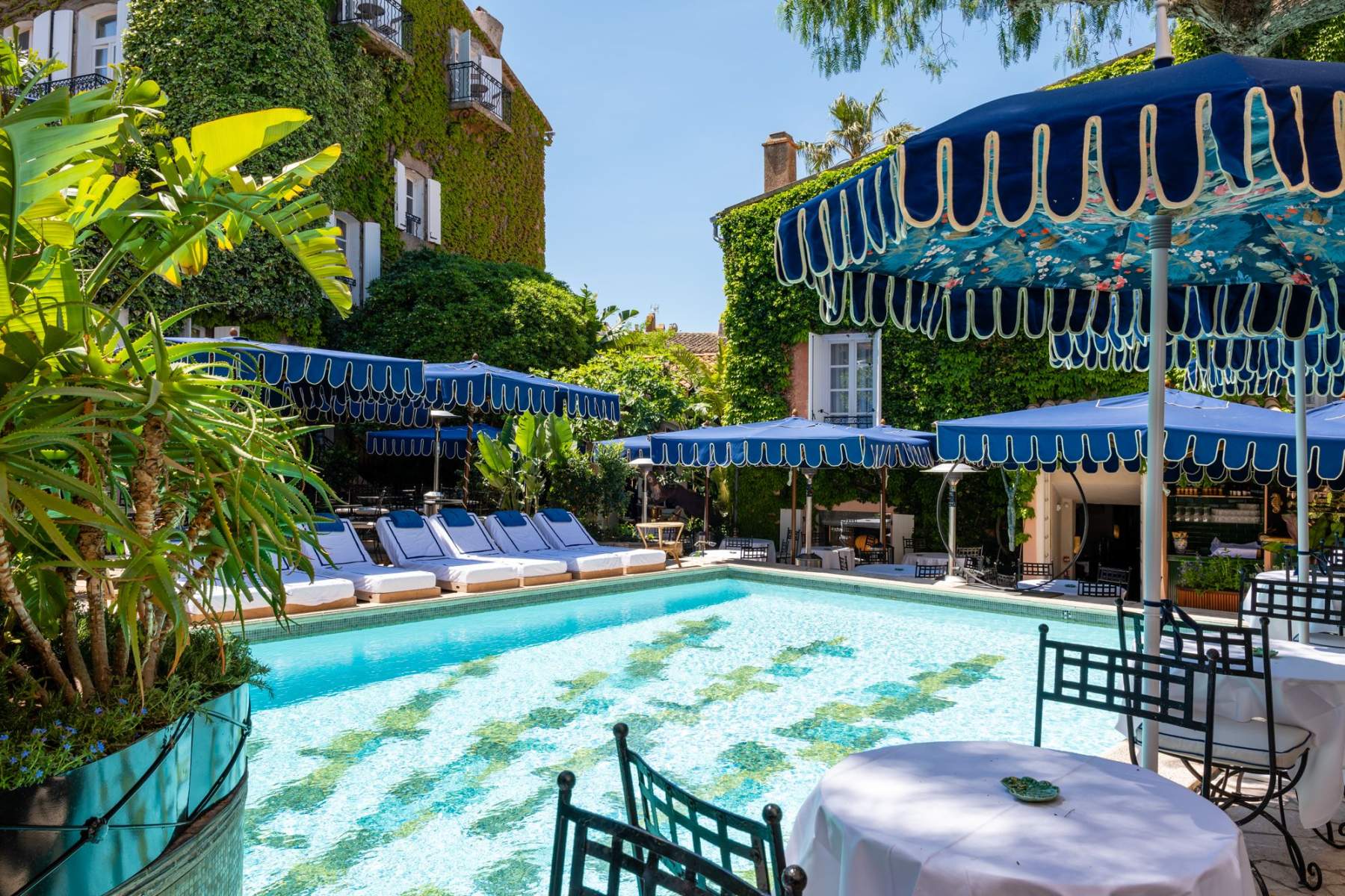 Poolside scene with turquoise water, white loungers under blue canopies and ivy-covered walls in a sunny resort setting.
