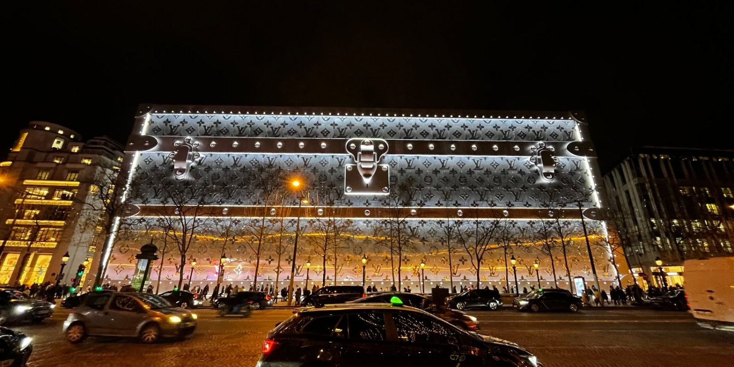 Le 103-111 Champs-Élysées habillé d’une malle Louis Vuitton géante illuminée, la nuit, sur l’avenue.