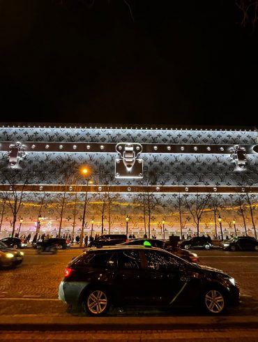 Le 103-111 Champs-Élysées habillé d’une malle Louis Vuitton géante illuminée, la nuit, sur l’avenue.
