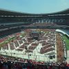 Large stadium filled with spectators in red, daytime event on the field with a central stage and rigging setup.