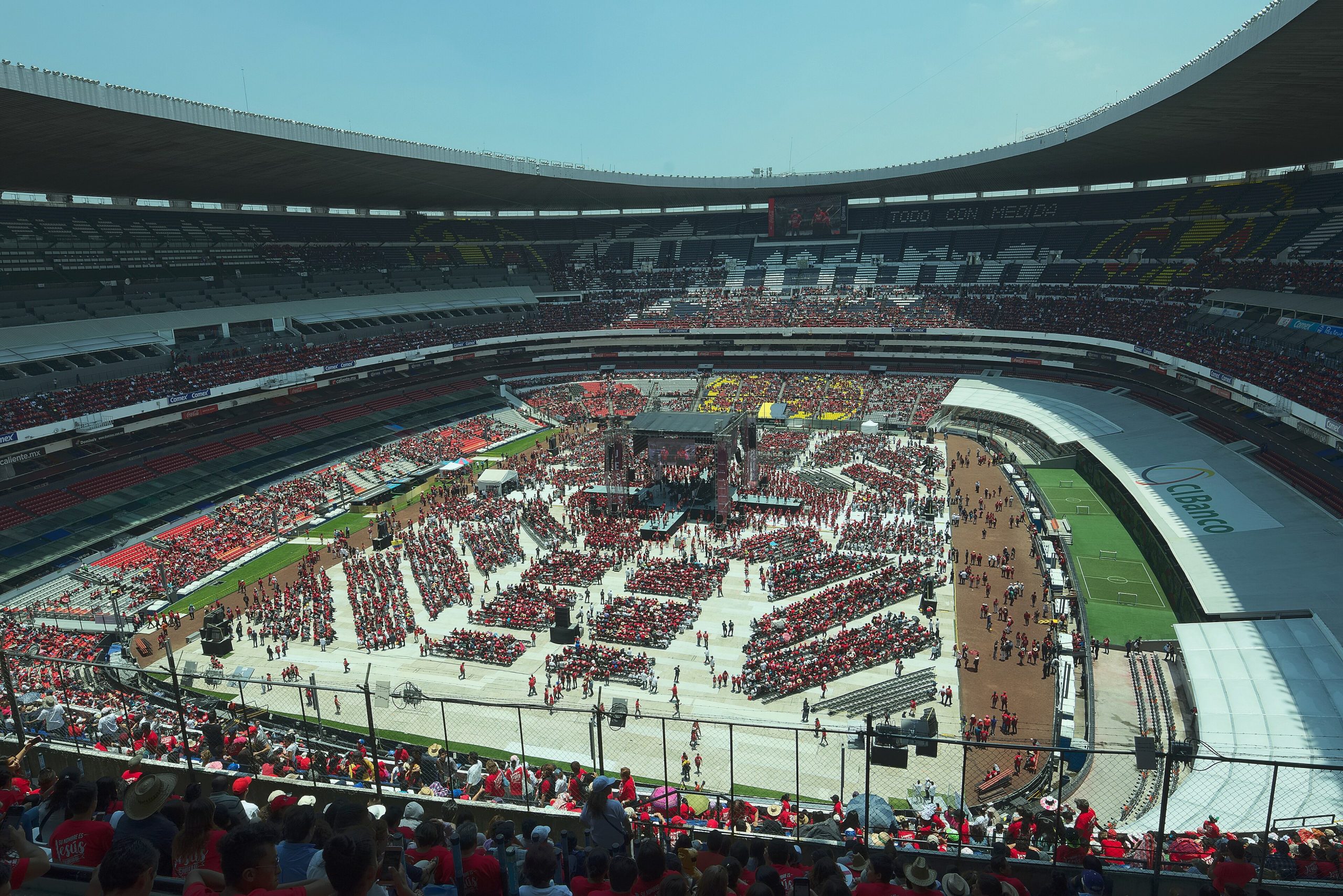 Large stadium filled with spectators in red, daytime event on the field with a central stage and rigging setup.