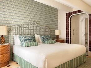 Elegant hotel bedroom featuring a large white bed with green-striped upholstered headboard and patterned throw pillows; two round wooden nightstands with lamps flank the bed.