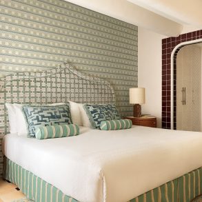 Elegant hotel bedroom featuring a large white bed with green-striped upholstered headboard and patterned throw pillows; two round wooden nightstands with lamps flank the bed.