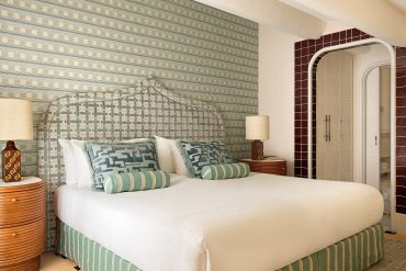 Elegant hotel bedroom featuring a large white bed with green-striped upholstered headboard and patterned throw pillows; two round wooden nightstands with lamps flank the bed.
