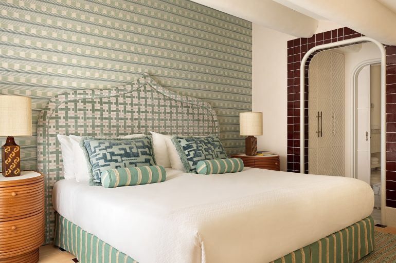 Elegant hotel bedroom featuring a large white bed with green-striped upholstered headboard and patterned throw pillows; two round wooden nightstands with lamps flank the bed.
