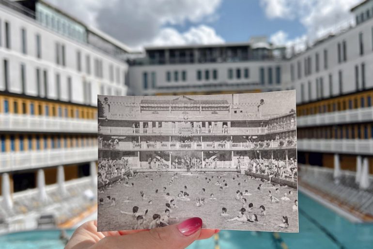 Person holding a black-and-white photo of a crowded outdoor event in front of a modern building courtyard pool area