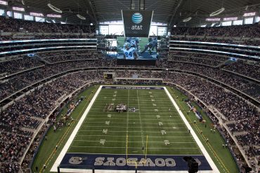 A packed football stadium with a massive video screen hanging over the field showing players, and the Cowboys end zone visible at the near end of the field.