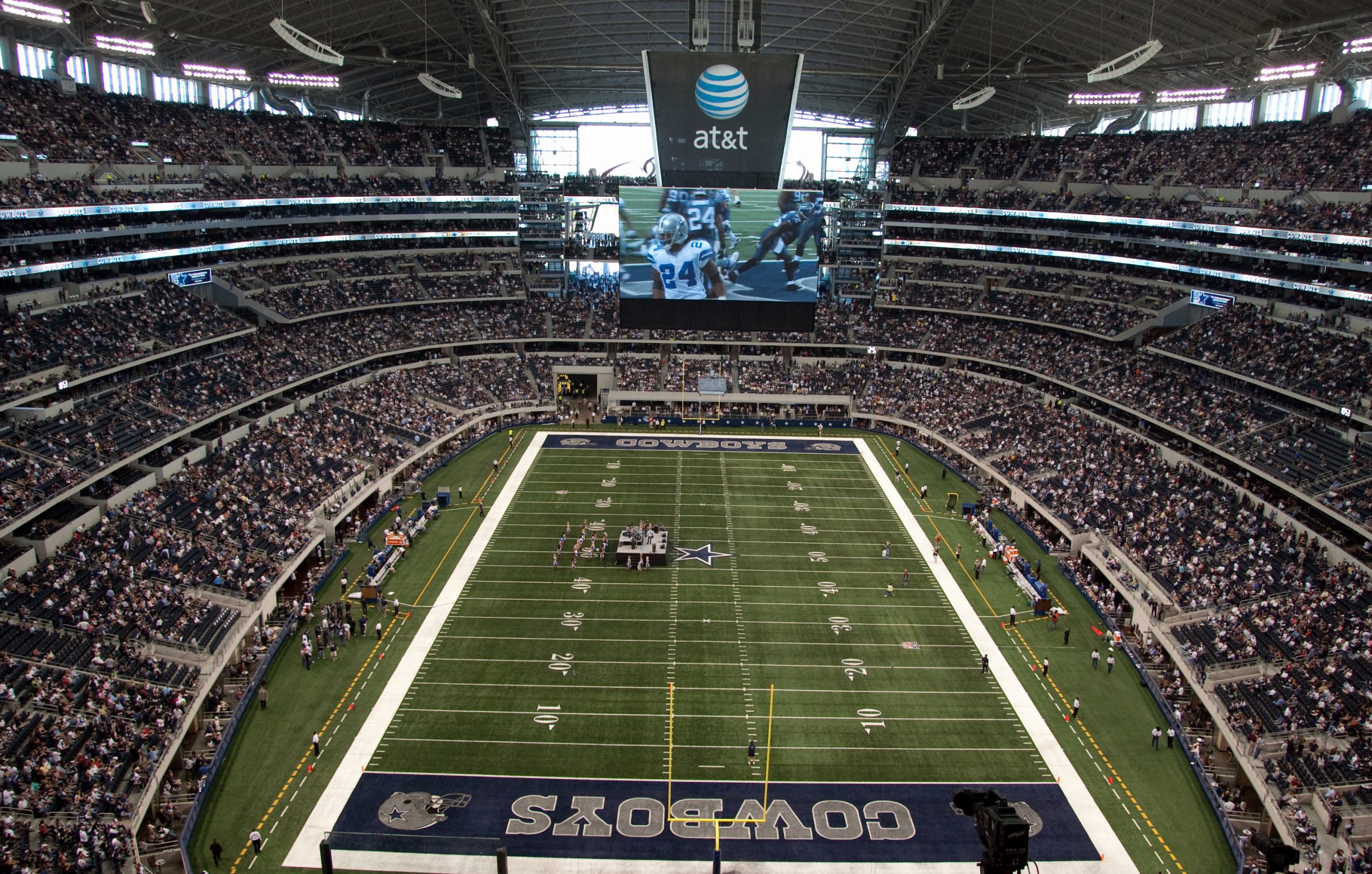 A packed football stadium with a massive video screen hanging over the field showing players, and the Cowboys end zone visible at the near end of the field.