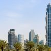 Dubai skyline with the Burj Khalifa on the left, blue glass skyscrapers on the right, and palm trees in the foreground under a clear sky.