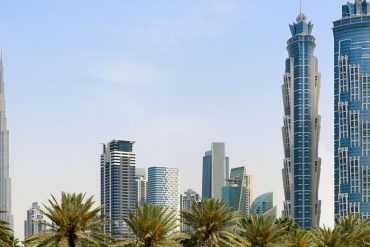 Dubai skyline with the Burj Khalifa on the left, blue glass skyscrapers on the right, and palm trees in the foreground under a clear sky.