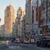 Wide city street lined with tall buildings on both sides, cars in traffic and traffic lights at dusk in Madrid-like downtown.