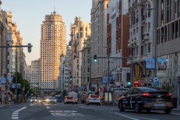 Wide city street lined with tall buildings on both sides, cars in traffic and traffic lights at dusk in Madrid-like downtown.
