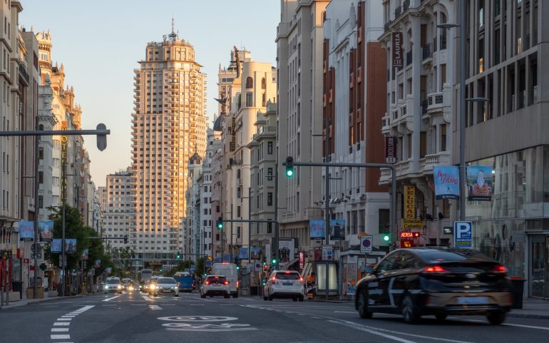 Wide city street lined with tall buildings on both sides, cars in traffic and traffic lights at dusk in Madrid-like downtown.