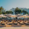 Beachfront lounge area with striped sunbeds under white umbrellas, Martinez hotel visible in the background.