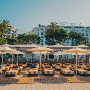 Beachfront lounge area with striped sunbeds under white umbrellas, Martinez hotel visible in the background.