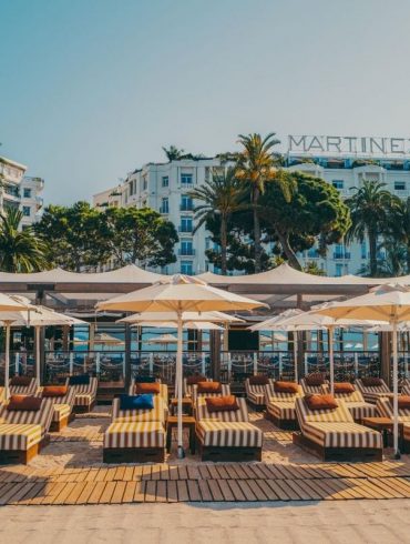 Beachfront lounge area with striped sunbeds under white umbrellas, Martinez hotel visible in the background.