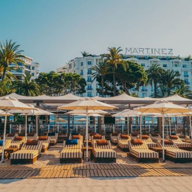 Beachfront lounge area with striped sunbeds under white umbrellas, Martinez hotel visible in the background.