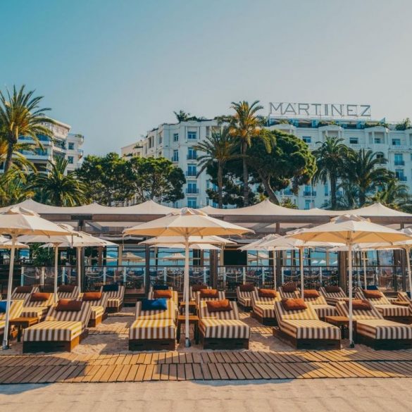 Beachfront lounge area with striped sunbeds under white umbrellas, Martinez hotel visible in the background.