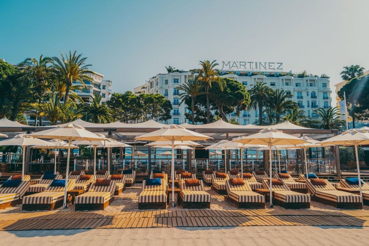 Beachfront lounge area with striped sunbeds under white umbrellas, Martinez hotel visible in the background.