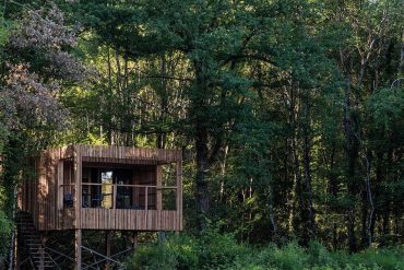 Wooden treehouse on stilts among a dense green forest, with a small balcony and chairs visible inside the cabin.