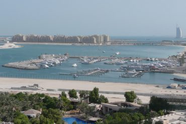 Harbor filled with boats at Dubai Marina, high-rise apartments along the shore, with the Burj Al Arab visible in the distance under a clear blue sky.