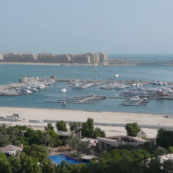 Harbor filled with boats at Dubai Marina, high-rise apartments along the shore, with the Burj Al Arab visible in the distance under a clear blue sky.